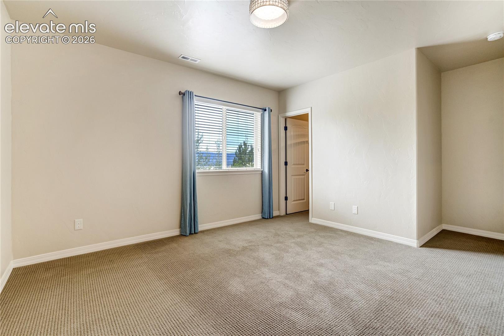 This neutral-toned bedroom features wall-to-wall carpeting and light-colored walls, creating a clean and versatile living space. A single window with blue curtains allows natural light to enter, while a doorway leads to an adjacent closet area. The room is illuminated by a central flush-mount light fixture, offering a simple and functional layout.