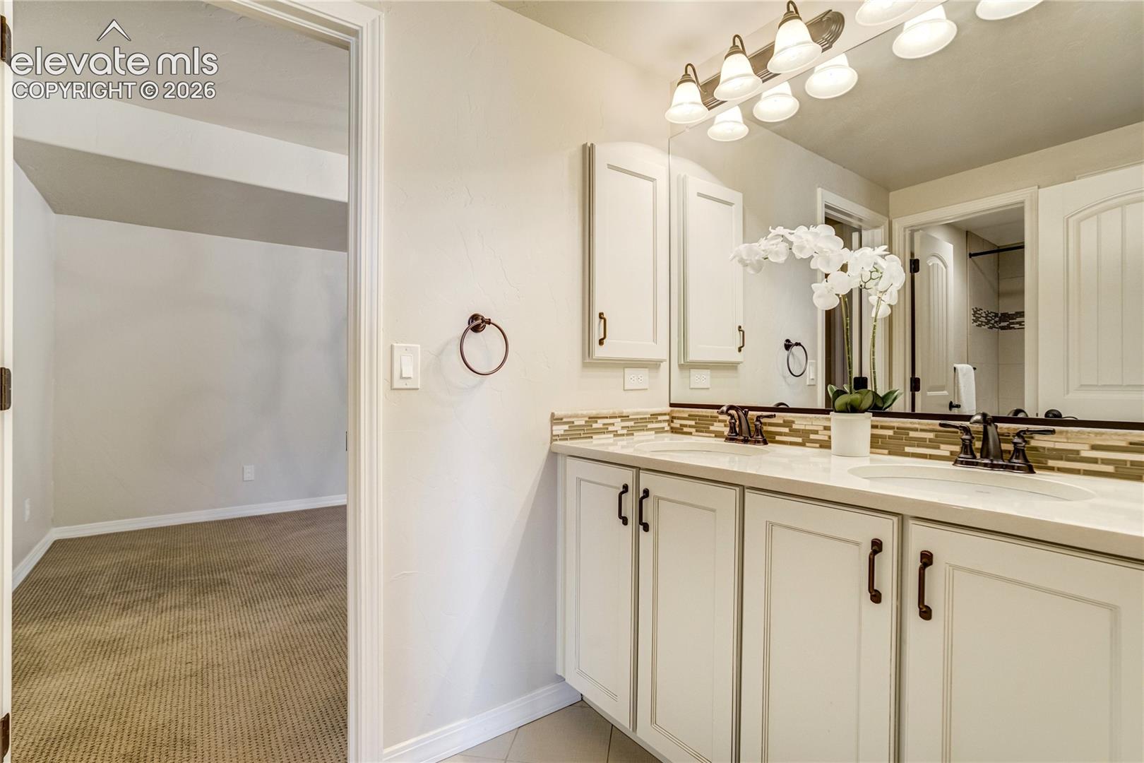 This bathroom features a double vanity with light-colored cabinetry, dark hardware, and a neutral countertop. A large mirror spans the wall above the vanity, complemented by a multi-light fixture and a decorative tile backsplash. The perspective looks into the bathroom from an adjacent carpeted room, highlighting the transition between spaces.