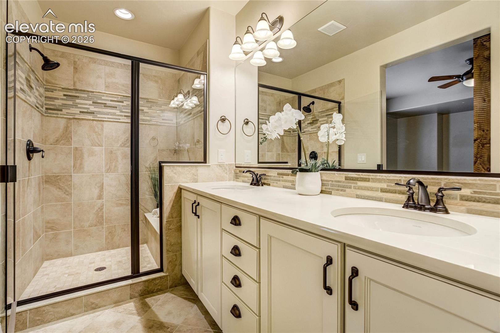 This primary bathroom features a double vanity with light-colored cabinetry, dark hardware, and a light-toned countertop. To the left, a walk-in shower is enclosed by a dark-framed glass door and finished with neutral-toned wall and floor tiles. A large mirror spans the wall above the vanity, reflecting the space and enhancing the bright, clean aesthetic of the room.