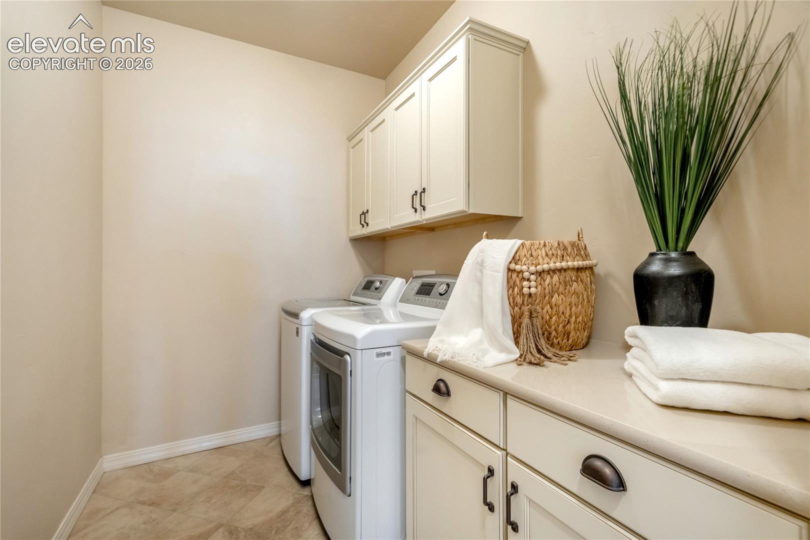 This functional laundry room features a clean, neutral aesthetic with light-toned cabinetry and matching countertops. The space is equipped with a side-by-side washer and dryer set, complemented by a woven basket, a decorative plant, and neatly folded white towels on the counter. The room is well-lit and organized, offering a practical and tidy workspace for household chores.