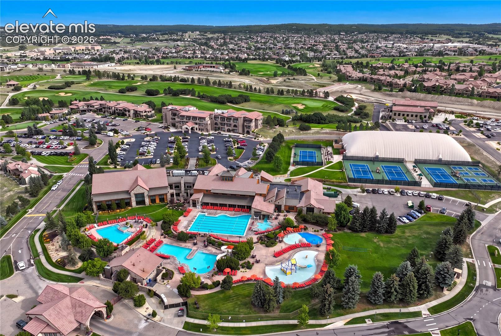 This high-angle aerial view captures a sprawling community recreation center featuring an expansive pool complex with multiple swimming areas, a splash pad, and lounge seating. Adjacent to the pools are several tennis courts, including an indoor facility under a large white dome, all set against a backdrop of a manicured golf course and residential neighborhood. The architecture of the main clubhouse building is traditional with pitched roofs, creating a cohesive and inviting atmosphere for community members.