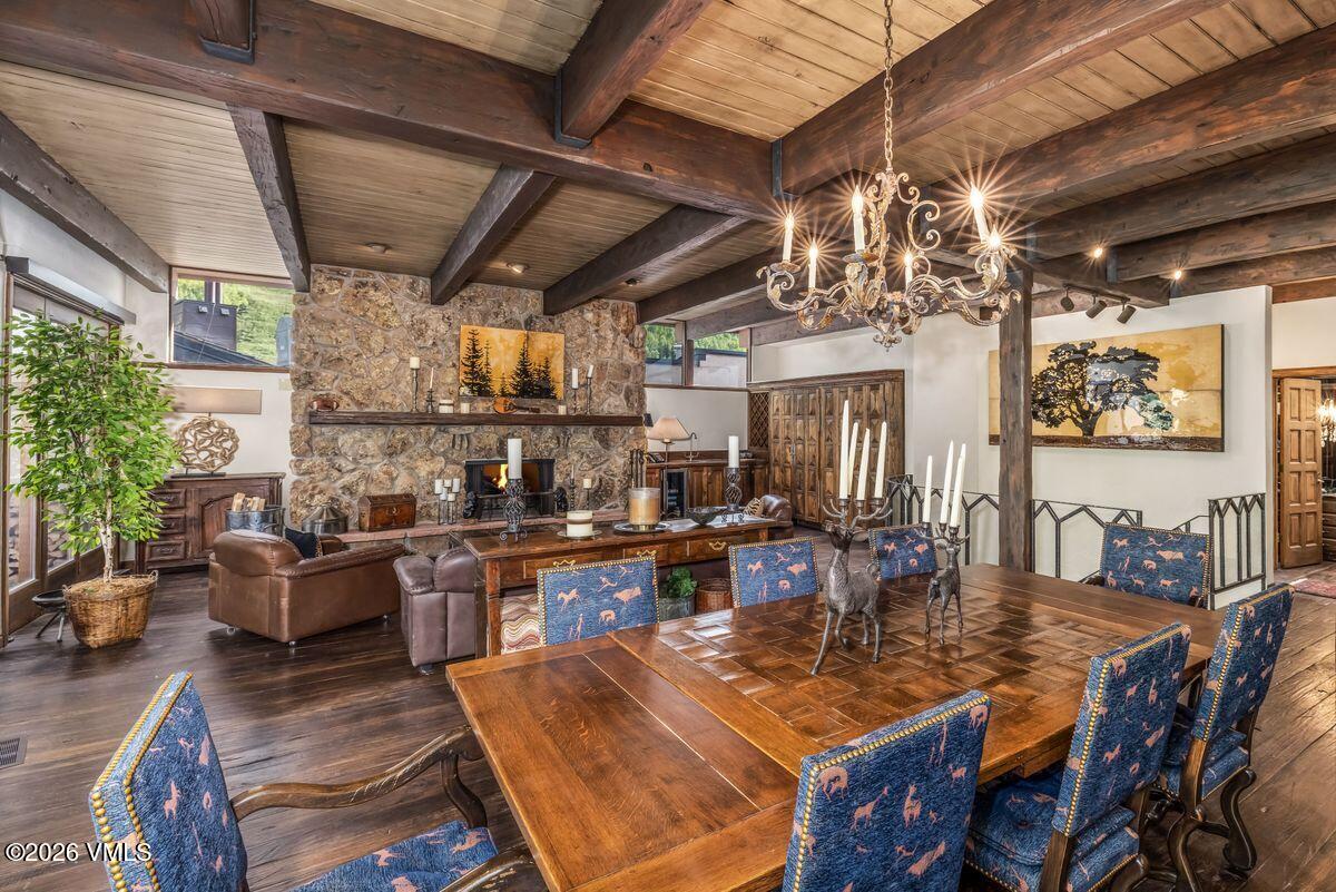 This rustic dining room features a large wooden table surrounded by chairs upholstered in a blue patterned fabric, set beneath an ornate metal chandelier. The space flows into a living area anchored by a stone-clad chimney breast and a wooden mantel, with exposed timber beams across the ceiling. The warm, inviting atmosphere is enhanced by dark wood flooring and a large potted plant near the windows, creating a cohesive mountain-lodge aesthetic.