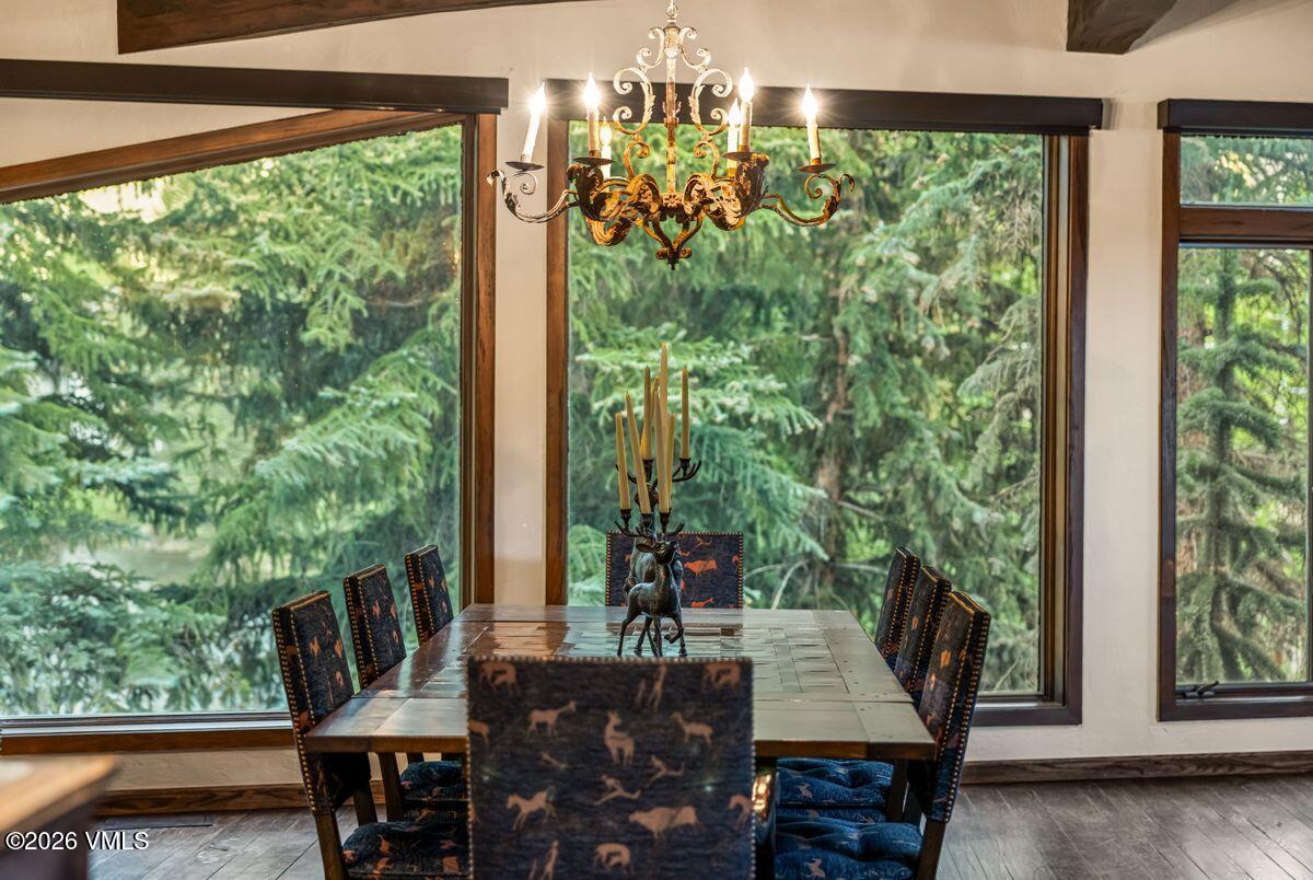 This dining room features a rustic wooden table surrounded by chairs upholstered in a dark fabric with a whimsical animal print pattern. A large, ornate metal chandelier hangs above the table, which is centered in front of expansive windows that provide a panoramic view of the surrounding forest. The room is framed by dark wood trim, creating a cozy, lodge-inspired atmosphere that emphasizes the connection to the natural landscape outside.