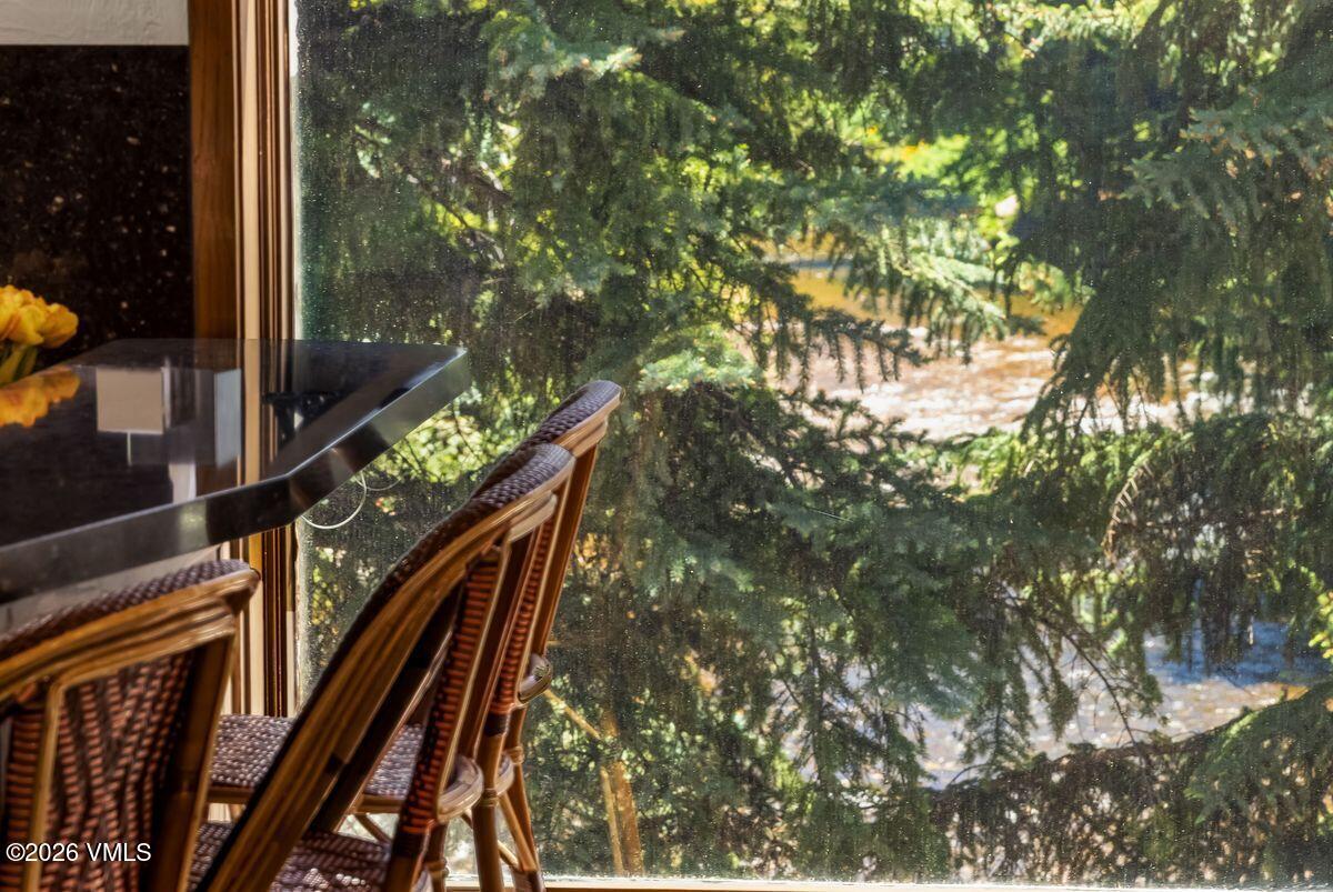 This interior shot captures a close-up view of a dark, polished countertop surface paired with woven-style chairs, positioned directly against a large window. Through the glass, a lush, dense forest scene provides a serene and natural backdrop, suggesting a peaceful, secluded setting. The perspective is intimate, focusing on the contrast between the sleek indoor furniture and the organic, textured greenery outside.