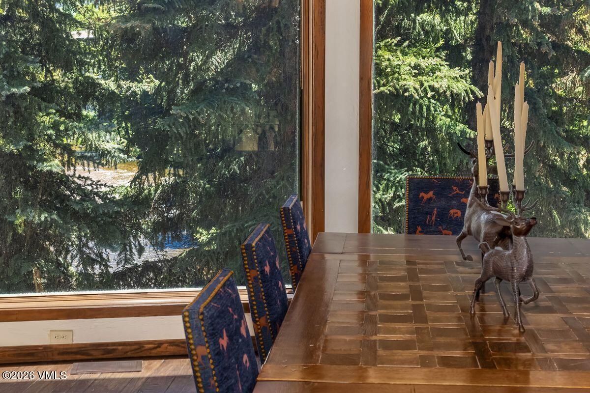 This dining area features a large, square wooden table with a distinctive woven-pattern top, surrounded by chairs upholstered in a dark blue fabric with a repeating animal motif. Large windows provide an expansive view of a lush, wooded forest and a nearby stream, bringing the natural environment into the living space. A decorative metal sculpture of deer holding candles serves as a centerpiece on the table, enhancing the rustic, nature-inspired aesthetic of the room.