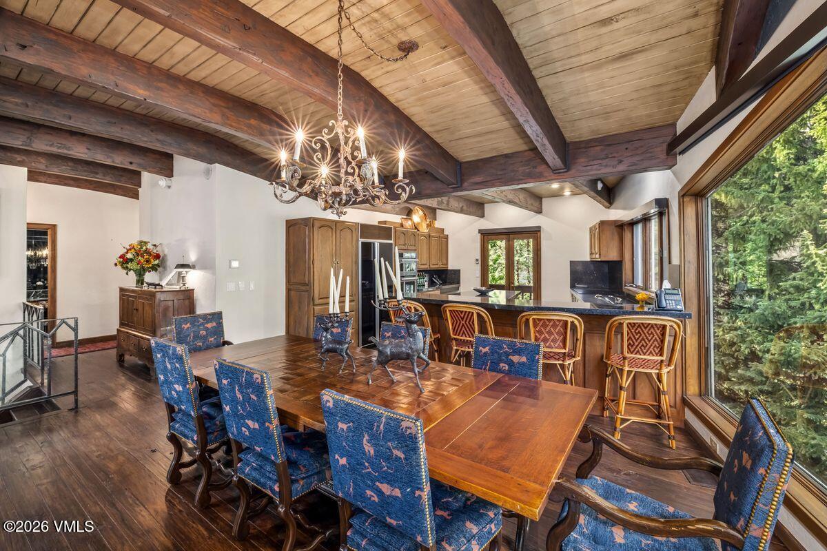 This open-concept dining area features a large wooden table surrounded by chairs upholstered in a blue fabric with a wildlife-inspired pattern. The space is defined by exposed wooden ceiling beams and a rustic chandelier, creating a warm, lodge-like atmosphere. Large windows along the right wall provide an expansive view of the surrounding forest, while the adjacent kitchen area includes a breakfast bar with matching stools.