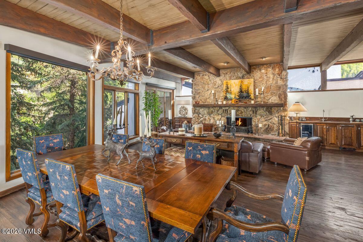 This rustic dining room features a large wooden table surrounded by chairs upholstered in a blue patterned fabric, set beneath a decorative chandelier. The space is defined by exposed wooden ceiling beams and a prominent stone-clad chimney breast housing a fireplace, which anchors the room. Large windows provide expansive views of the surrounding forest, creating a warm and inviting atmosphere that blends indoor comfort with natural elements.