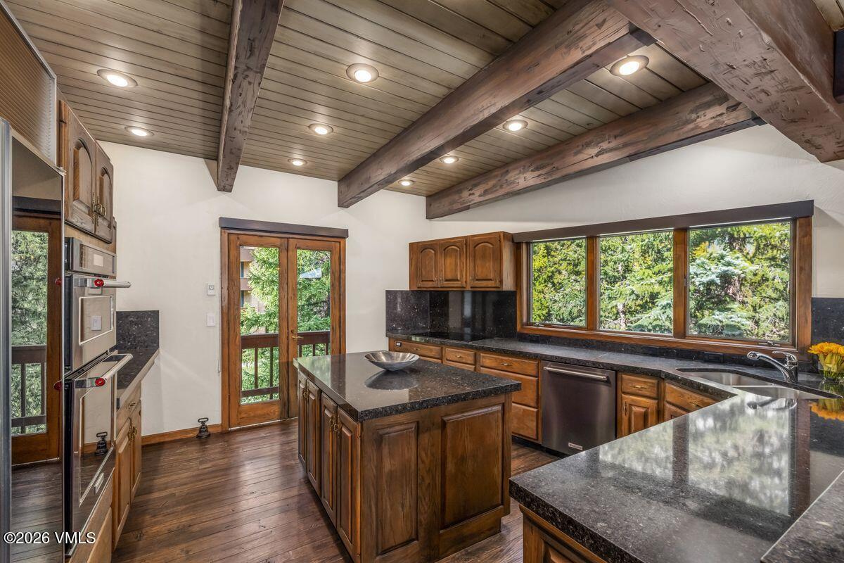 This rustic-inspired kitchen features dark stone countertops, warm wood cabinetry, and a central island that serves as a focal point. The space is defined by a vaulted ceiling with exposed wooden beams and recessed lighting, creating a cozy, mountain-lodge atmosphere. Large windows provide a scenic view of the surrounding trees, while a glass-paned door offers direct access to an outdoor deck.