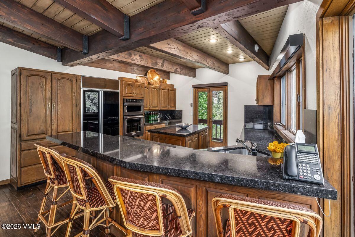 This rustic-style kitchen features dark wood cabinetry, exposed ceiling beams, and a large island with a dark stone countertop. A breakfast bar with three woven chairs overlooks the workspace, which includes a built-in oven, a sink, and a view through glass doors to the outdoors. The space is illuminated by recessed lighting and maintains a warm, traditional aesthetic.