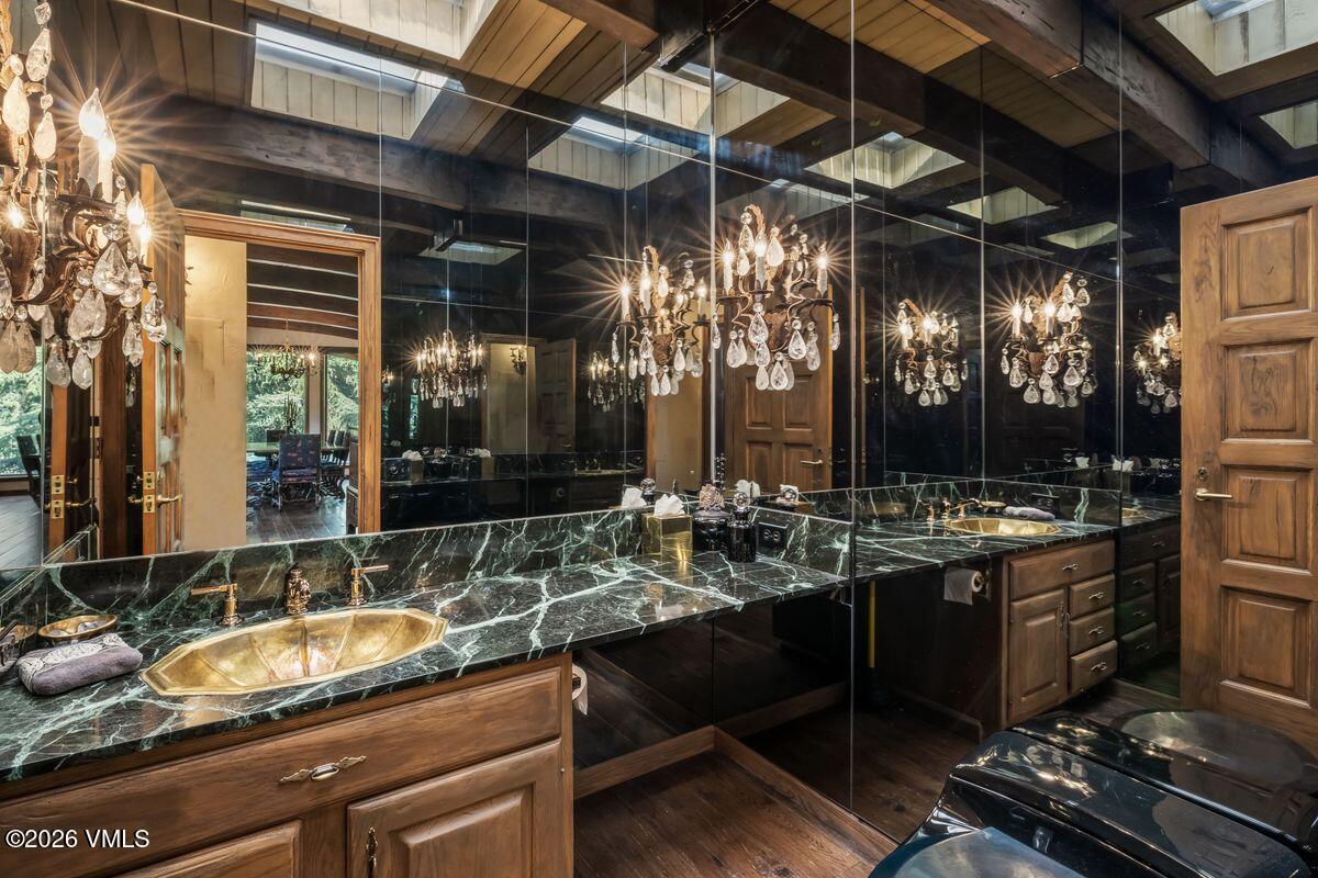 This bathroom features a striking vanity with a dark, heavily veined stone countertop and a metallic, oval-shaped drop-in sink. A large, wall-to-wall mirror reflects the room, including a decorative crystal chandelier and exposed wooden ceiling beams. The space is finished with warm-toned wood cabinetry and dark wood flooring, creating a dramatic and opulent aesthetic.