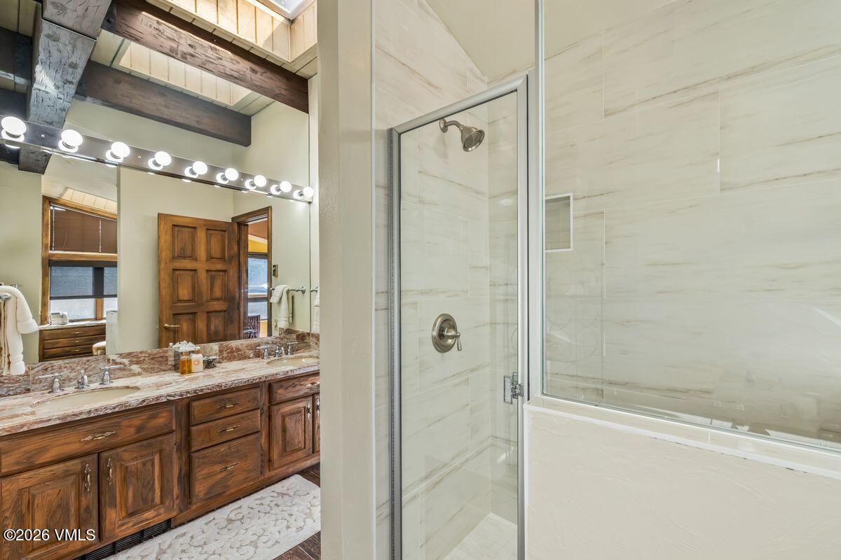This bathroom features a double vanity with a warm-toned wood cabinet and a patterned stone countertop, reflected in a large mirror spanning the wall. To the right, a glass-enclosed shower stall is finished with light-colored, large-format wall tiles. The space is accented by exposed dark wood ceiling beams, creating a rustic yet refined aesthetic.