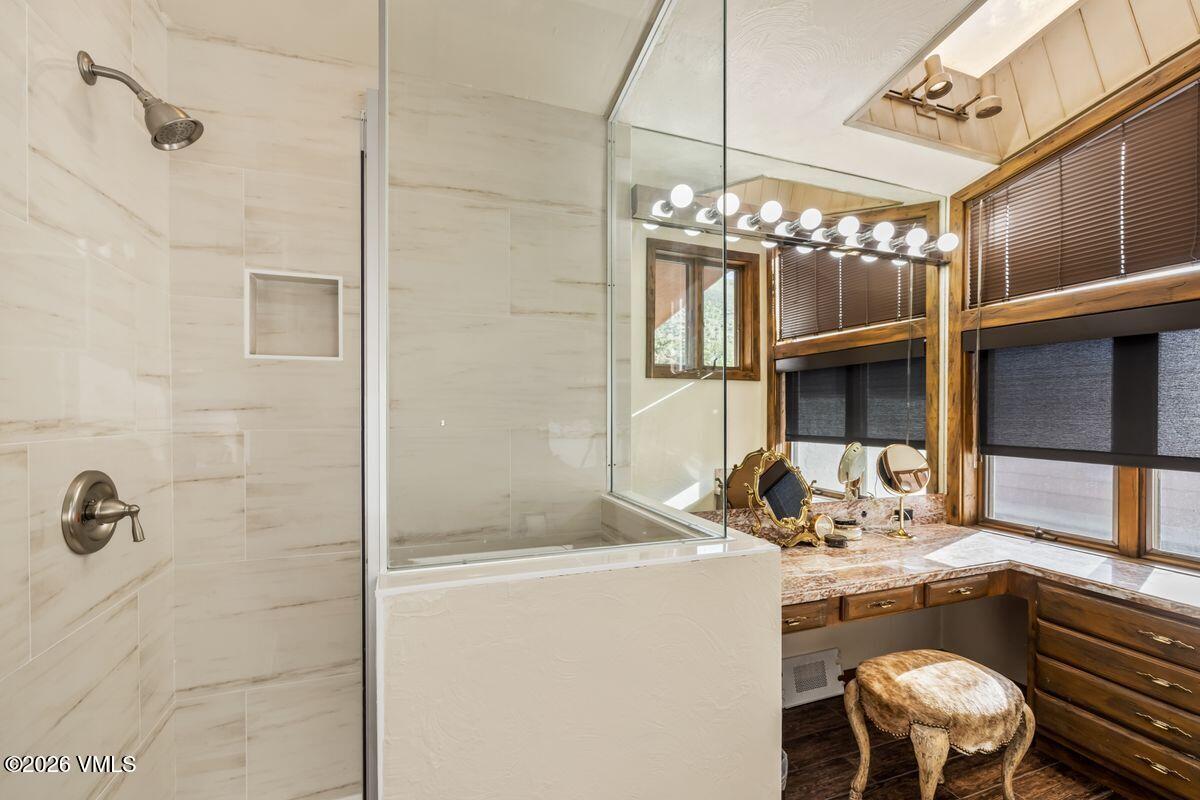 This primary bathroom features a glass-enclosed shower with light-toned, large-format wall tiles and a built-in recessed niche. Adjacent to the shower is a vanity area with a stone countertop, a multi-bulb light fixture, and a stool with a patterned seat. The space is brightened by large windows with dark blinds, creating a functional and well-lit grooming area.