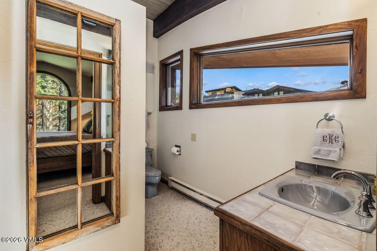 This bathroom features a vanity with a tiled countertop and a drop-in sink, accented by a towel hanging on a circular ring. A large, multi-pane mirror on the left wall reflects a glimpse into an adjacent bedroom, while a horizontal window above the vanity provides natural light and a view of the outdoors. The space is finished with neutral walls and a textured floor, creating a functional and compact layout.