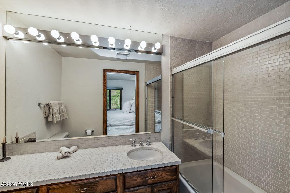 This bathroom features a vanity with a tiled countertop and a dark wood cabinet, paired with a large wall-to-wall mirror and a multi-bulb light fixture. To the right, a glass-enclosed shower stall is finished with neutral-toned square wall tiles. The perspective captures the vanity area and the shower, with a reflection in the mirror showing a glimpse of an adjacent bedroom.
