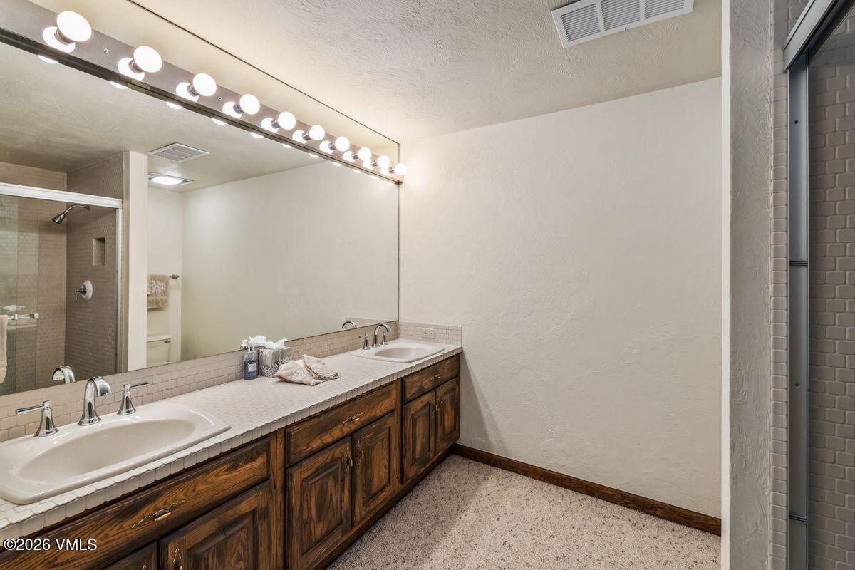 This bathroom features a long, dual-sink vanity with dark wood cabinetry and a tiled countertop that extends into a backsplash. A large, wall-to-wall mirror reflects the space, including the shower area and the lighting fixture above. The room is finished with neutral walls and carpeted flooring, providing a functional and traditional layout.
