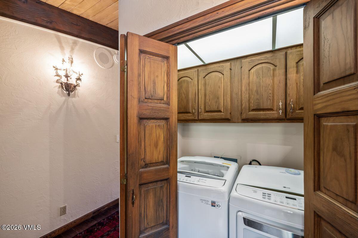 This laundry room is accessed through a set of double wooden doors, revealing a functional space equipped with a side-by-side washer and dryer. Above the appliances, wooden cabinetry provides overhead storage, while a recessed light fixture illuminates the area. To the left, a decorative wall sconce adds a touch of character to the adjacent hallway wall, which features a textured finish and a glimpse of a patterned rug.