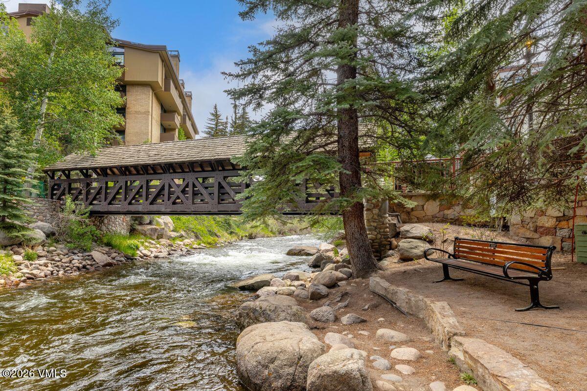 This scenic outdoor area features a rustic covered wooden bridge spanning a rushing mountain stream, surrounded by lush green trees and rocky banks. A wooden park bench sits on a stone-paved path in the foreground, offering a peaceful spot to enjoy the natural surroundings. The perspective is at eye level, capturing the tranquil atmosphere of a community park or common area adjacent to a multi-story residential building.