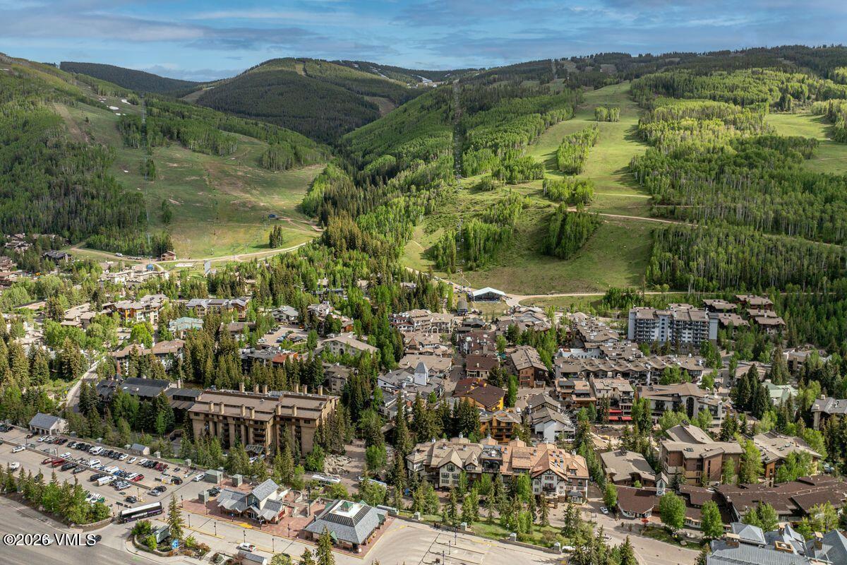 This high-angle aerial view captures a sprawling mountain resort village nestled at the base of a lush, green ski mountain. The scene features a dense collection of multi-story lodges and residential buildings with varied rooflines, surrounded by mature pine and aspen trees. The perspective provides a comprehensive look at the community layout, highlighting the proximity to the ski slopes and the surrounding natural landscape.