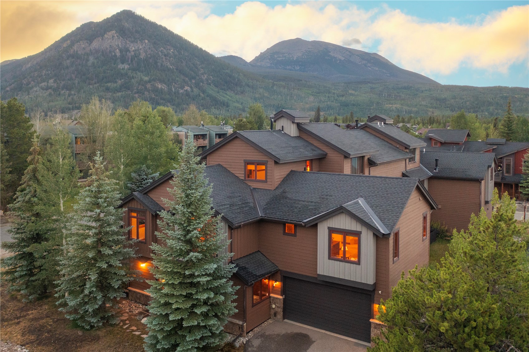 This elevated perspective captures a multi-story mountain home nestled against a backdrop of dramatic peaks and evergreen forests. The structure features a blend of brown wood siding, dark shingled roofing, and a prominent two-car garage, with warm interior lighting glowing through the windows. The setting conveys a cozy, rustic aesthetic typical of a high-altitude retreat.