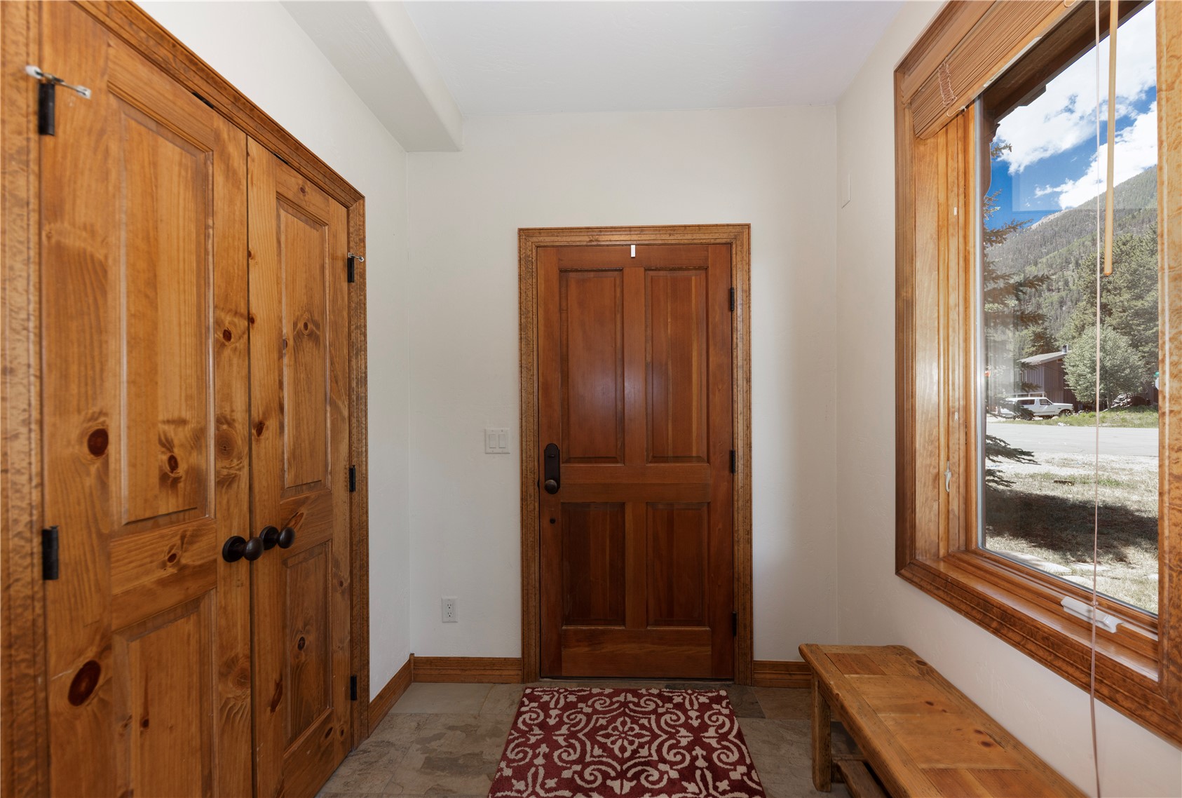 This inviting entryway features warm wood-toned doors and trim, creating a rustic and welcoming atmosphere. A patterned rug sits on the stone-tiled floor, while a wooden bench placed beneath a large window provides a functional seating area. The space is bright and airy, with a clear view through the window of the surrounding mountain landscape.