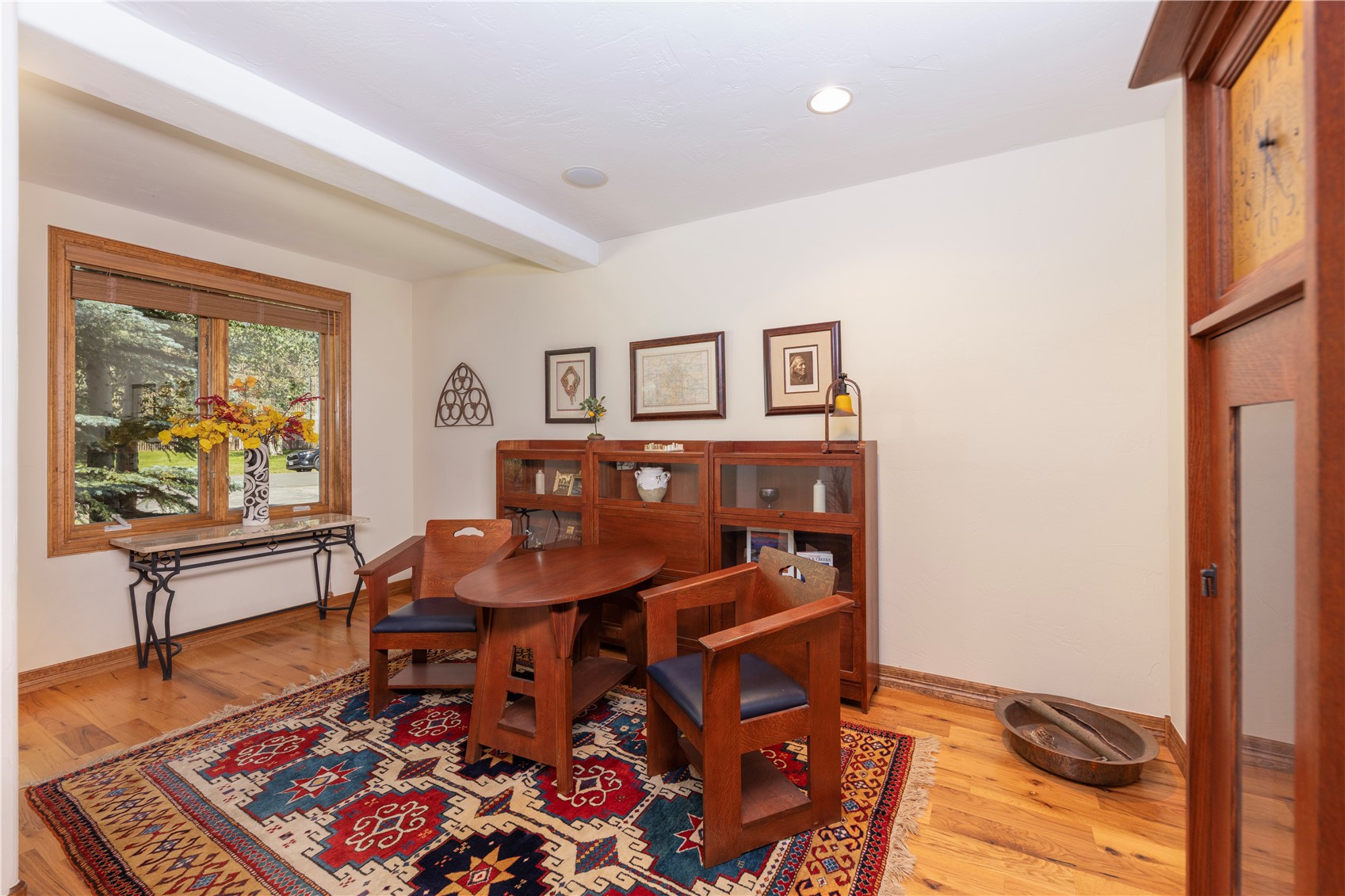 This cozy study features a warm, traditional aesthetic with rich wood furniture, including a small oval table paired with two matching chairs. A large, intricate patterned area rug anchors the space, while a wooden bookcase unit provides storage against the wall. The room is illuminated by recessed lighting and natural light streaming through a large window, creating a quiet and scholarly atmosphere.