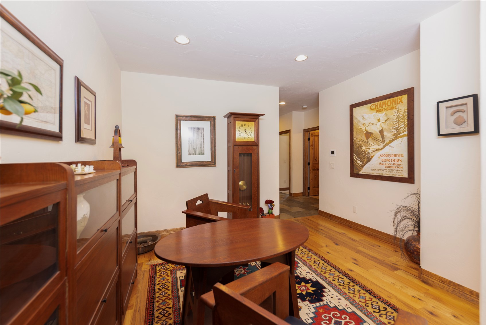 This cozy study or reading nook features warm wood flooring, a central oval table with matching chairs, and a tall grandfather clock standing against the wall. The space is decorated with framed artwork, including a vintage Chamonix ski poster, and a patterned area rug that adds texture to the room. A wooden cabinet with glass-front doors sits to the left, creating a classic, scholarly atmosphere.