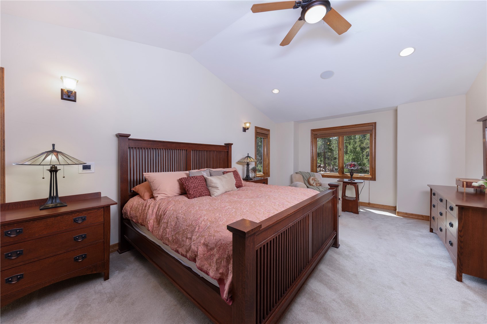This spacious primary bedroom features a large wooden bed frame with a slatted headboard and footboard, centered in the room. The space is accented by matching wooden dressers, a ceiling fan, and warm wall sconces, creating a cozy and traditional atmosphere. Large windows allow natural light to fill the room, offering a view of the surrounding trees.