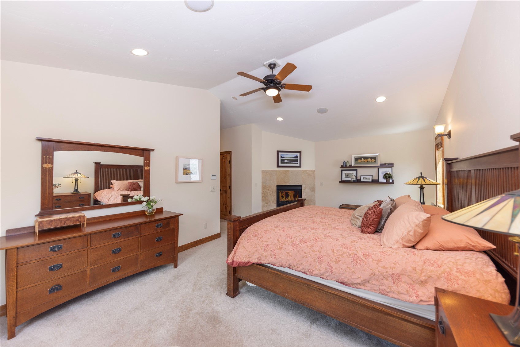 This spacious primary bedroom features a warm, traditional aesthetic with a large wooden bed frame and matching dresser. A stone-clad chimney breast houses a fireplace, creating a cozy focal point, while a wall-mounted shelf displays framed art and decor. The room is illuminated by a ceiling fan with integrated lighting and recessed lights, all set against a neutral color palette and carpeted flooring.