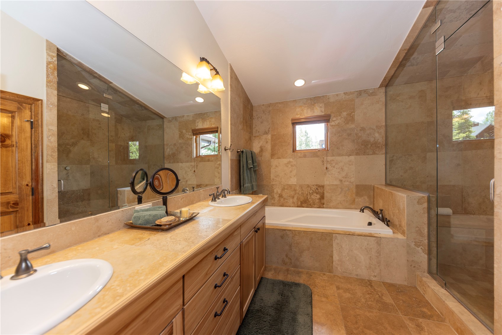 This primary bathroom features a dual-sink vanity with light wood cabinetry and a stone countertop, complemented by a large wall-to-wall mirror. The space is finished with warm-toned stone tile on the walls and floors, incorporating a built-in soaking tub and a separate glass-enclosed shower. The perspective captures the full length of the vanity and the integrated tub area, creating a cohesive and spa-like atmosphere.