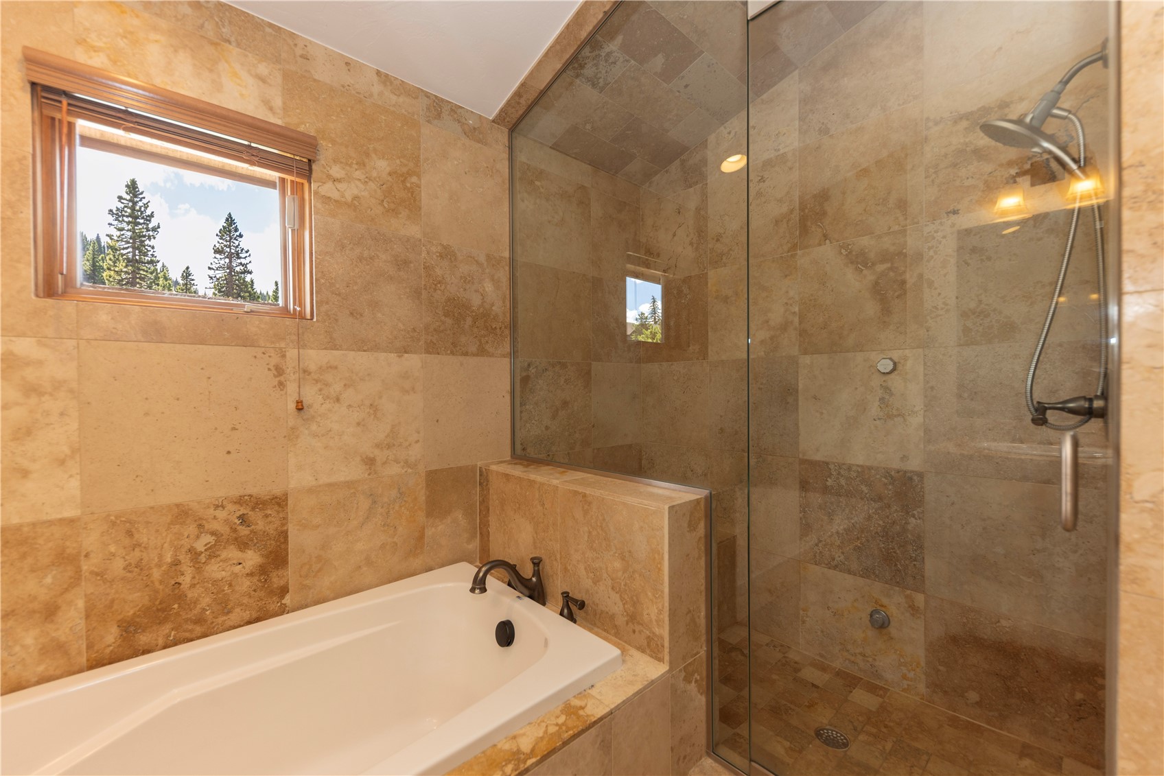 This bathroom features a combined soaking tub and walk-in shower area finished in warm-toned, textured stone tiles. A glass partition separates the shower from the tub, while a window above the tub provides natural light and a view of the surrounding trees. The space is designed with a cohesive, earthy aesthetic that emphasizes a spa-like atmosphere.