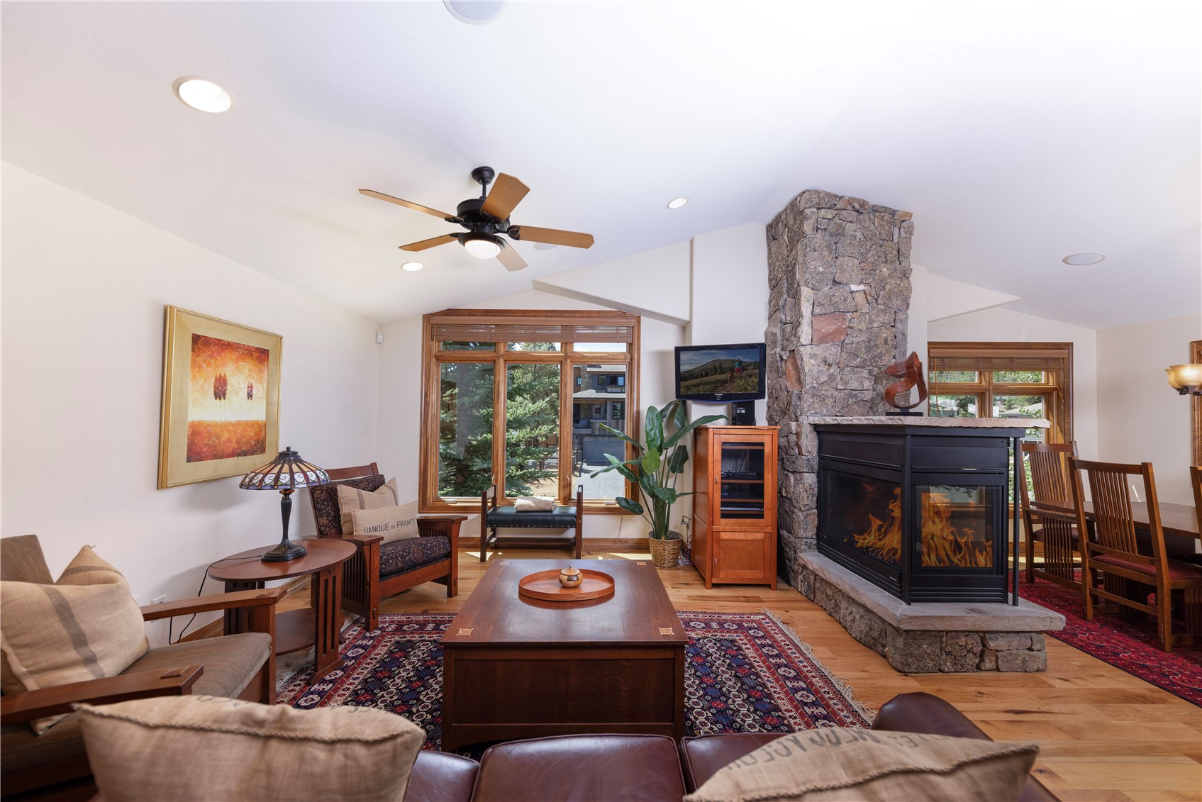 This inviting living room features a warm, rustic aesthetic with light wood flooring and a prominent stone-clad chimney breast housing a fireplace. The space is furnished with a leather sofa, a central wooden coffee table, and a patterned area rug, while large windows provide a view of the outdoors. A ceiling fan with integrated lighting centers the room, which flows seamlessly into an adjacent dining area.