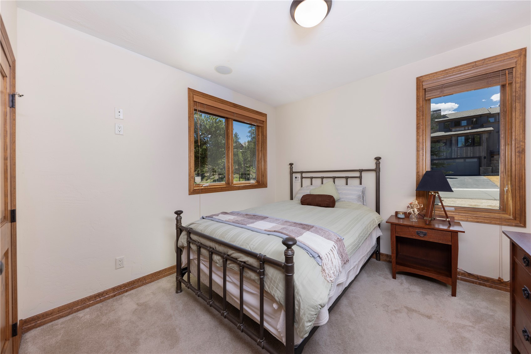 This cozy guest bedroom features a metal-framed bed dressed in light green bedding with a plaid throw blanket. The room is accented by warm wood-trimmed windows that provide natural light and views of the surrounding neighborhood. A small wooden nightstand with a black lamp sits beside the bed, completing the simple and inviting aesthetic of the space.