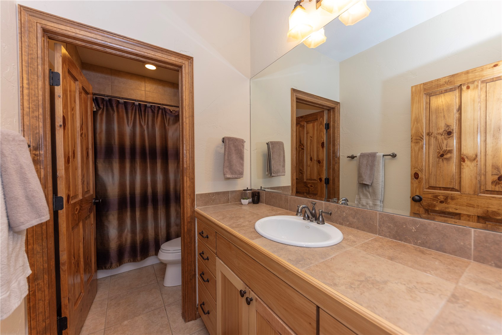 This bathroom features a vanity with a light wood finish, a neutral-toned stone countertop, and an oval undermount sink. A large wall-mounted mirror reflects the room, while a separate area houses a toilet and a shower with a dark patterned curtain. The space is accented by warm-toned wood trim on the doors and cabinetry, creating a cohesive and rustic aesthetic.