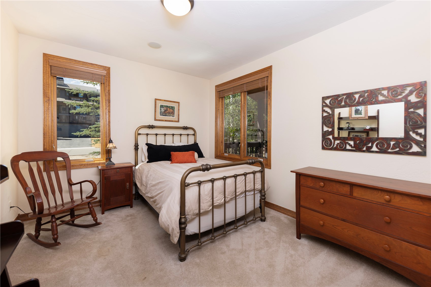 This cozy guest bedroom features a metal-framed bed centered between two windows with wood trim. The room is furnished with a wooden rocking chair, a bedside table with a small lamp, and a wooden dresser topped with a decorative mirror. The space is finished with neutral-toned walls and carpet, creating a warm and inviting atmosphere.
