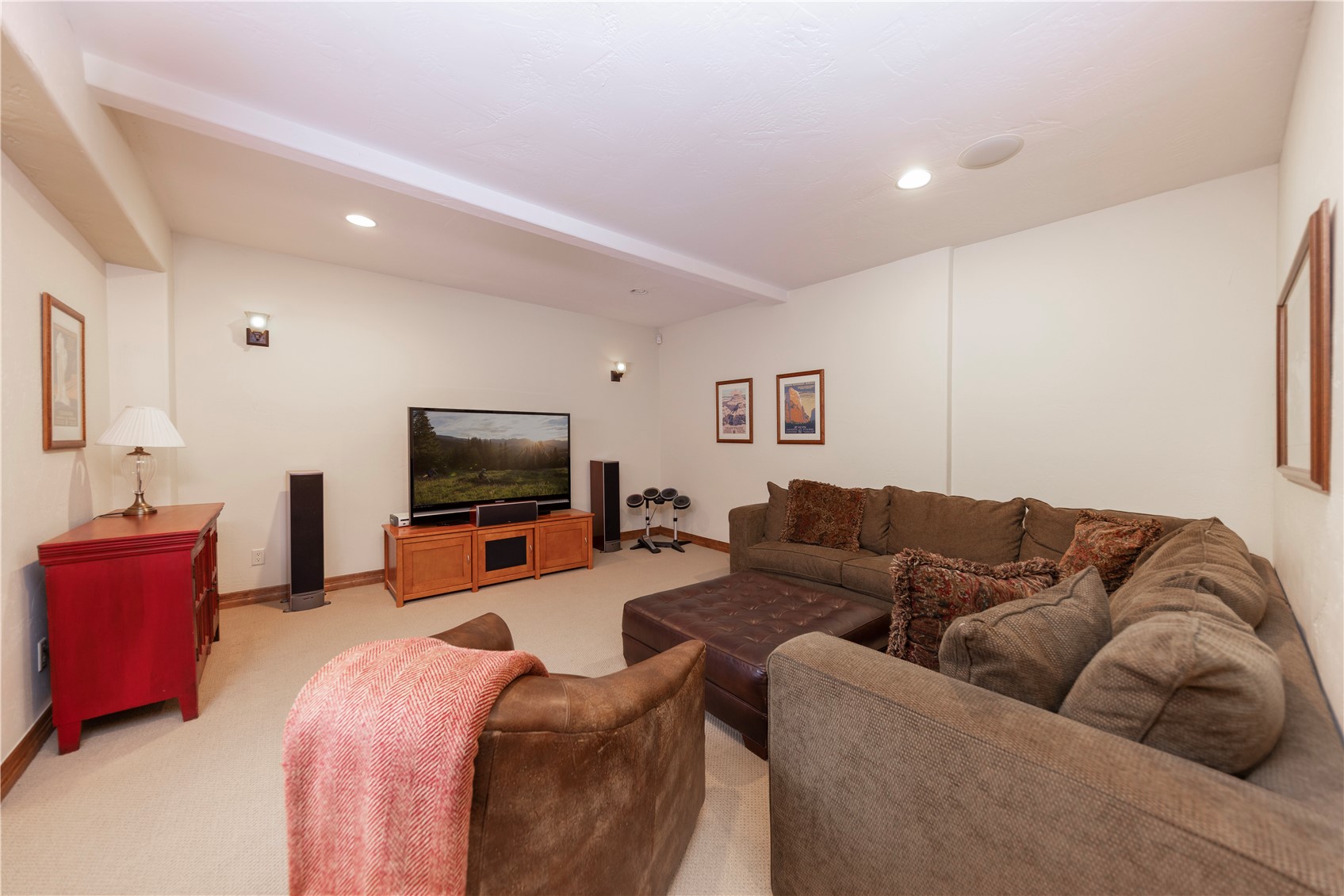 This cozy basement living area features a large, L-shaped sectional sofa paired with a leather ottoman and a comfortable armchair, creating an inviting space for relaxation. The room is equipped with a television on a wooden media console, flanked by floor-standing speakers, and decorated with framed artwork on the neutral-toned walls. Recessed lighting and wall sconces provide soft illumination, while a red cabinet adds a pop of color to the otherwise understated, carpeted space.