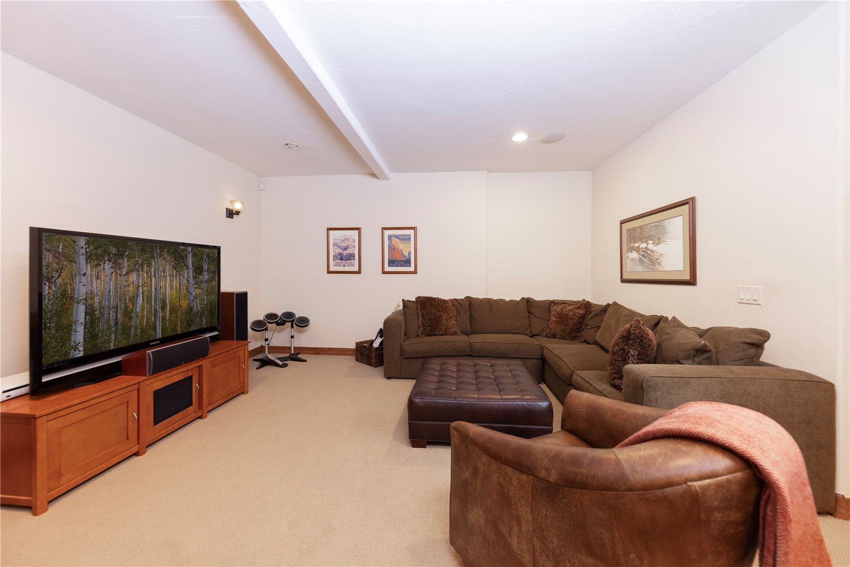 This spacious basement living room features a large sectional sofa paired with a leather ottoman and a comfortable armchair, creating a cozy seating area. A large television sits atop a wooden media console, complemented by a small electronic drum set in the corner. The room is finished with neutral carpeting and framed wall art, offering a relaxed and functional space for entertainment.