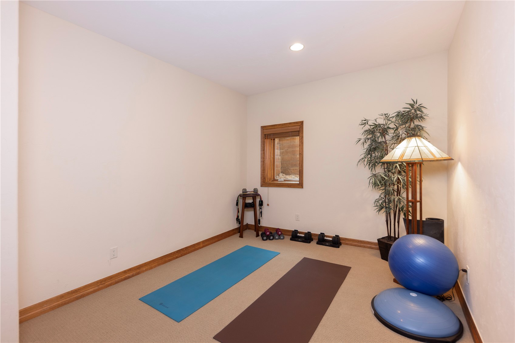 This home gym features a neutral color palette with beige carpeting and light-colored walls, creating a clean and functional workout space. The room is equipped with two yoga mats, a blue exercise ball, a balance trainer, and various hand weights, all arranged neatly on the floor. A small window with a wood frame provides natural light, while a decorative floor lamp and a potted plant add a touch of warmth to the corner.