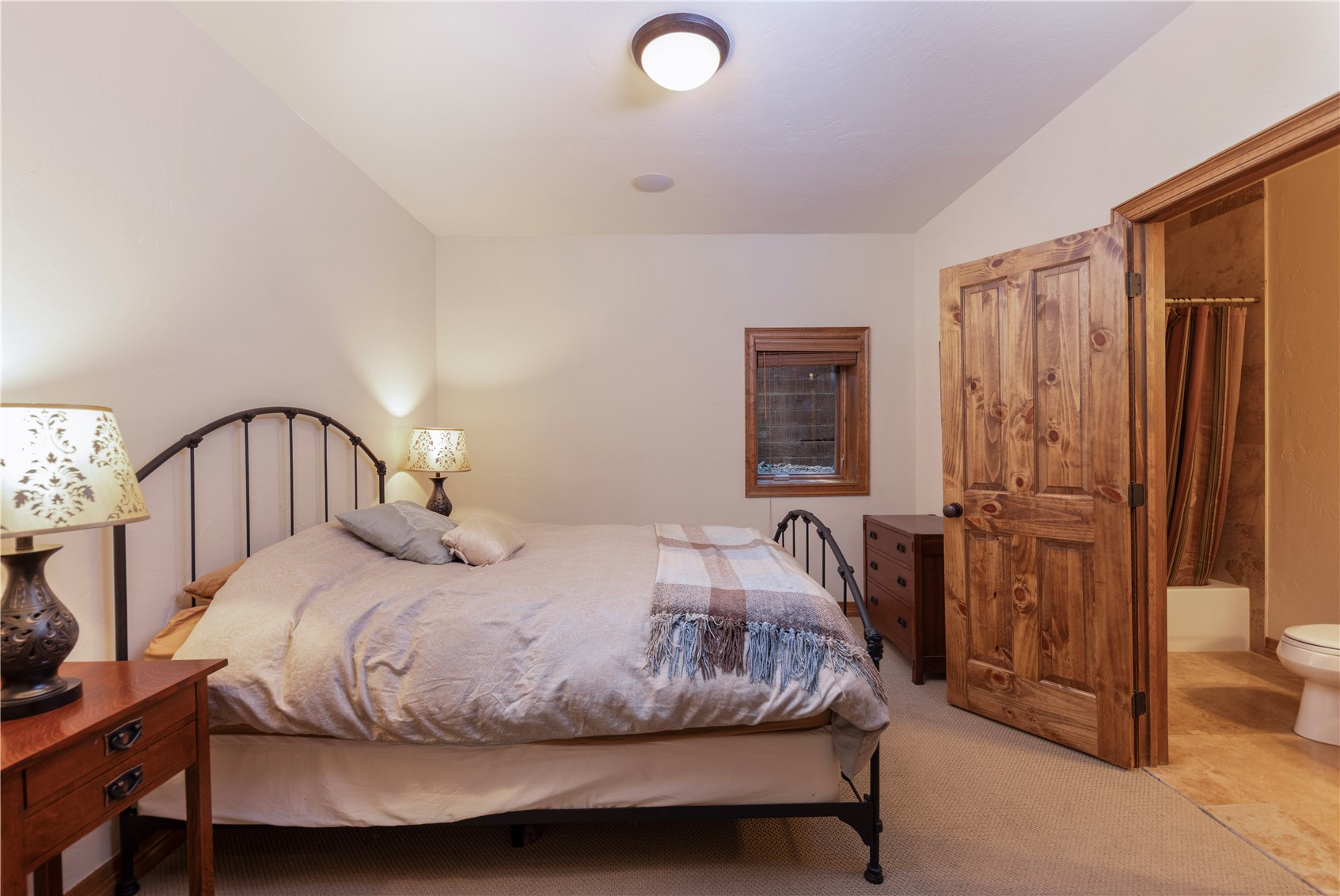 This cozy bedroom features a dark metal bed frame centered against a neutral wall, complemented by a wooden nightstand and a matching dresser. A small, wood-framed window provides natural light, while a rustic wooden door leads into an adjacent bathroom with a shower and toilet. The room is finished with neutral carpeting and a simple ceiling light fixture, creating a warm and inviting atmosphere.
