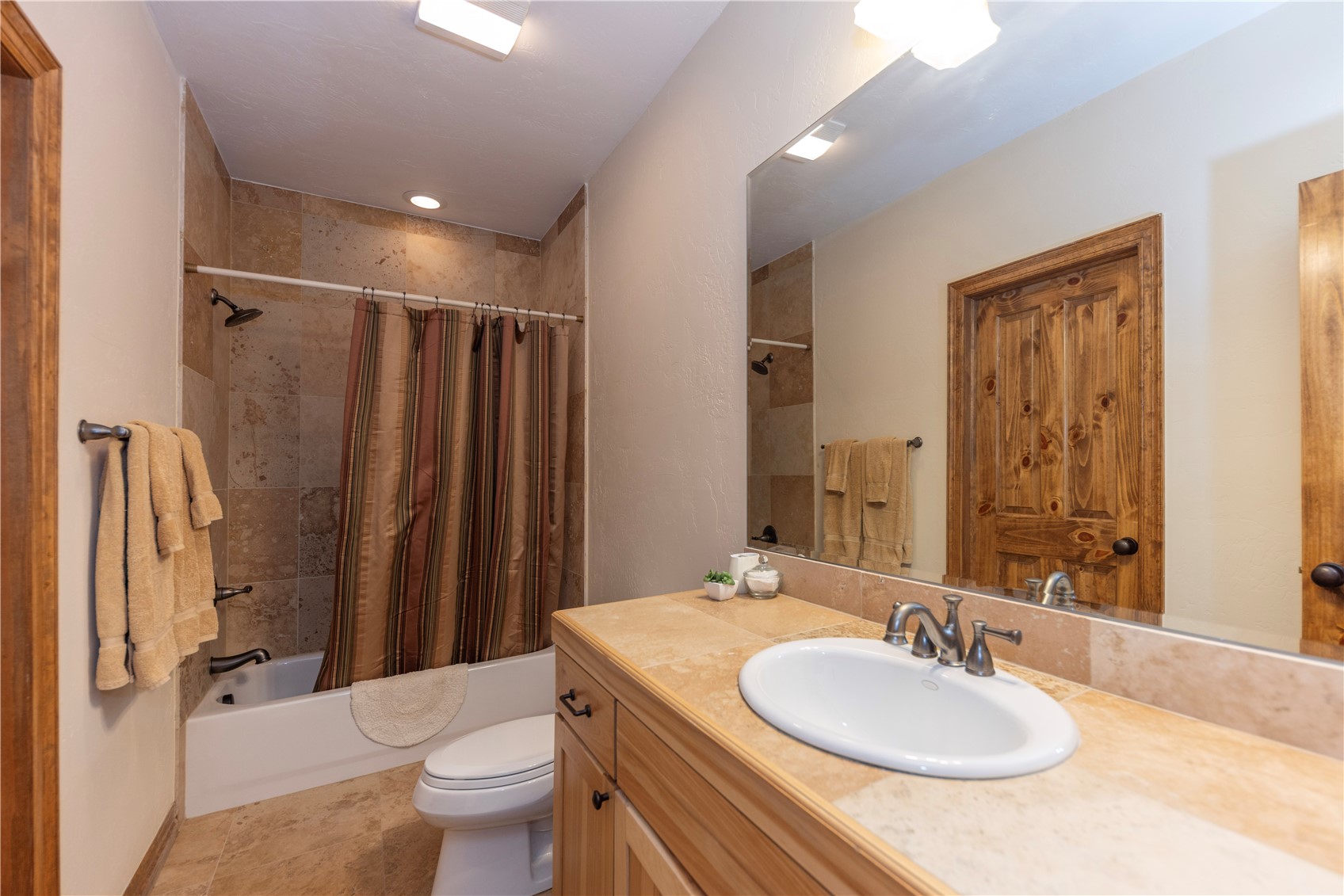 This bathroom features a vanity with a light-toned wood cabinet and a neutral stone countertop, paired with a large wall-mounted mirror. The shower-tub combination is enclosed by a striped curtain and finished with square stone tiles that extend to the ceiling. The room is completed by a white toilet and a matching stone-tiled floor, creating a cohesive and rustic aesthetic.