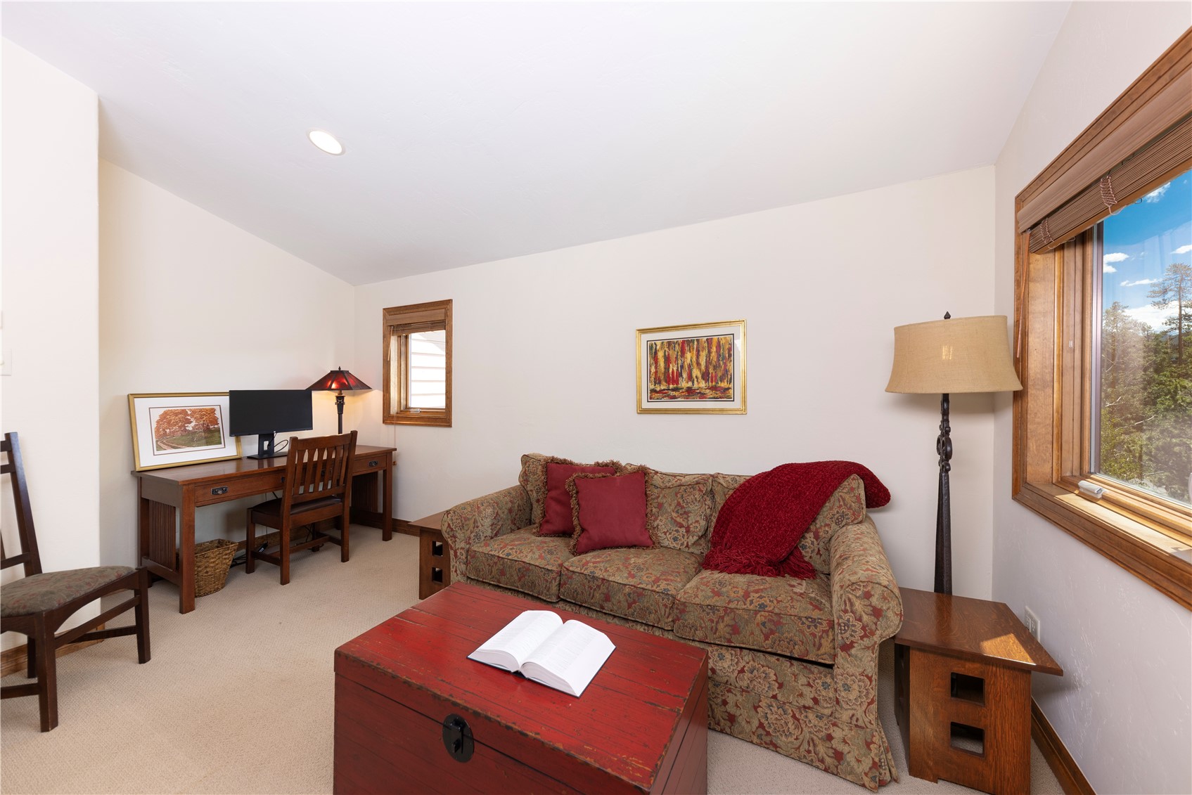 This versatile room functions as a cozy home office and sitting area, featuring a patterned sofa with red accent pillows and a matching throw blanket. A rustic red wooden chest serves as a coffee table in the center, while a wooden desk with a computer setup is positioned against the wall. The space is illuminated by a floor lamp and recessed lighting, with large windows providing natural light and a view of the surrounding trees.