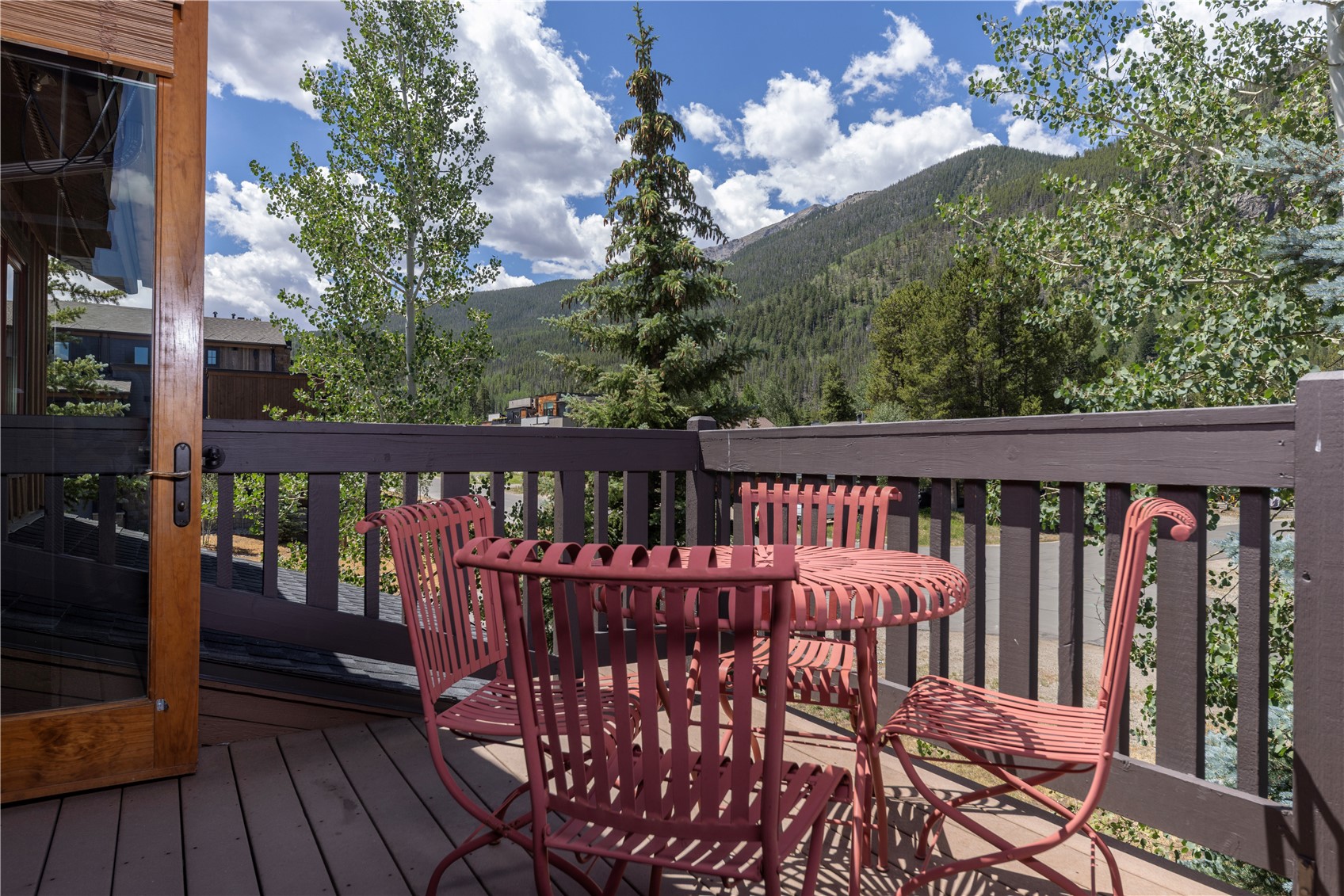 This elevated wooden deck features a set of red metal patio furniture, including a round table and four chairs, perfect for outdoor dining. The space is enclosed by a dark-painted wooden railing and offers a scenic, unobstructed view of a lush mountain landscape under a bright, cloudy sky. A glass-paneled door with a wooden frame provides direct access from the interior to this inviting outdoor living area.