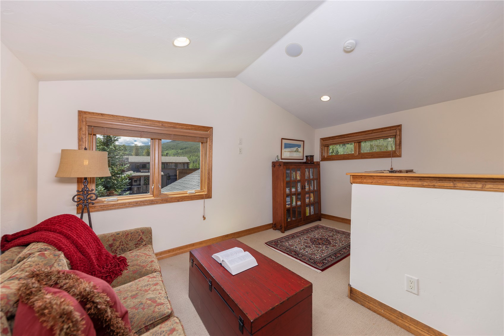 This cozy loft-style living area features a vaulted ceiling and a comfortable sofa adorned with a red throw blanket. A rustic red wooden chest serves as a coffee table, positioned in front of a window that offers a scenic view of the surrounding landscape. The space is completed by a wooden bookcase, a patterned area rug, and a floor lamp, creating a warm and inviting atmosphere.