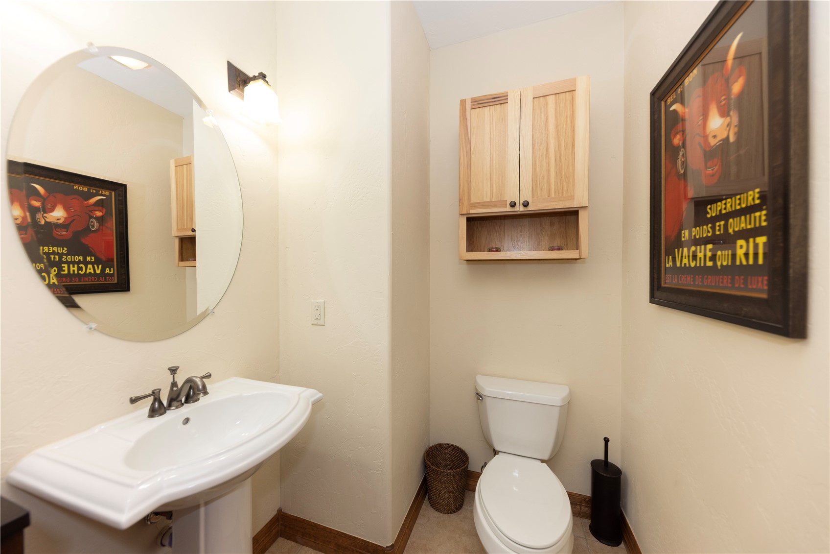 This compact powder room features a white pedestal sink paired with a round mirror and a wall-mounted light fixture. A light wood cabinet with open shelving is mounted above a standard white toilet, while a framed vintage-style advertisement adds a touch of character to the neutral-toned walls. The space is functional and well-lit, offering a clean and simple aesthetic.