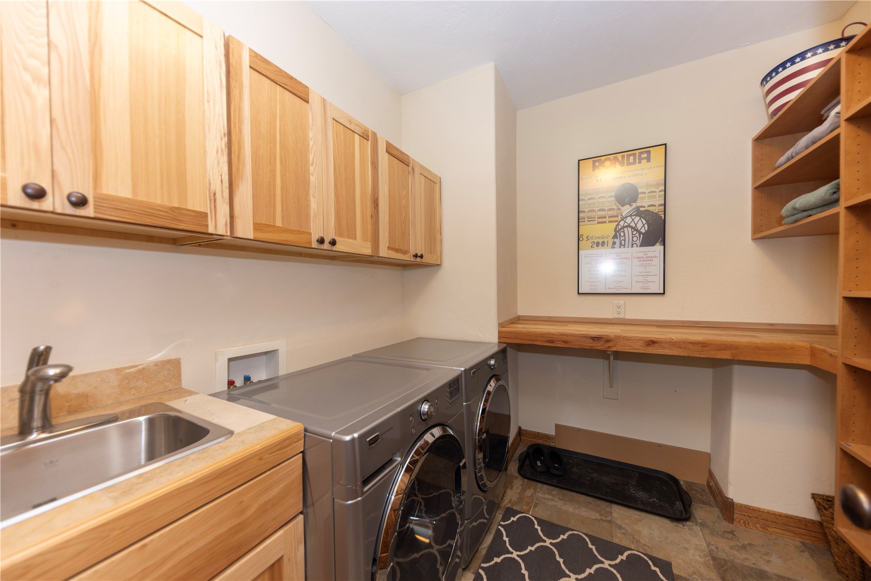 This functional laundry room features light wood cabinetry, a stainless steel sink, and a pair of silver front-loading appliances. A built-in wooden counter provides workspace, complemented by open shelving on the right and a decorative poster on the wall. The space is finished with neutral walls and patterned tile flooring, creating a practical and organized utility area.