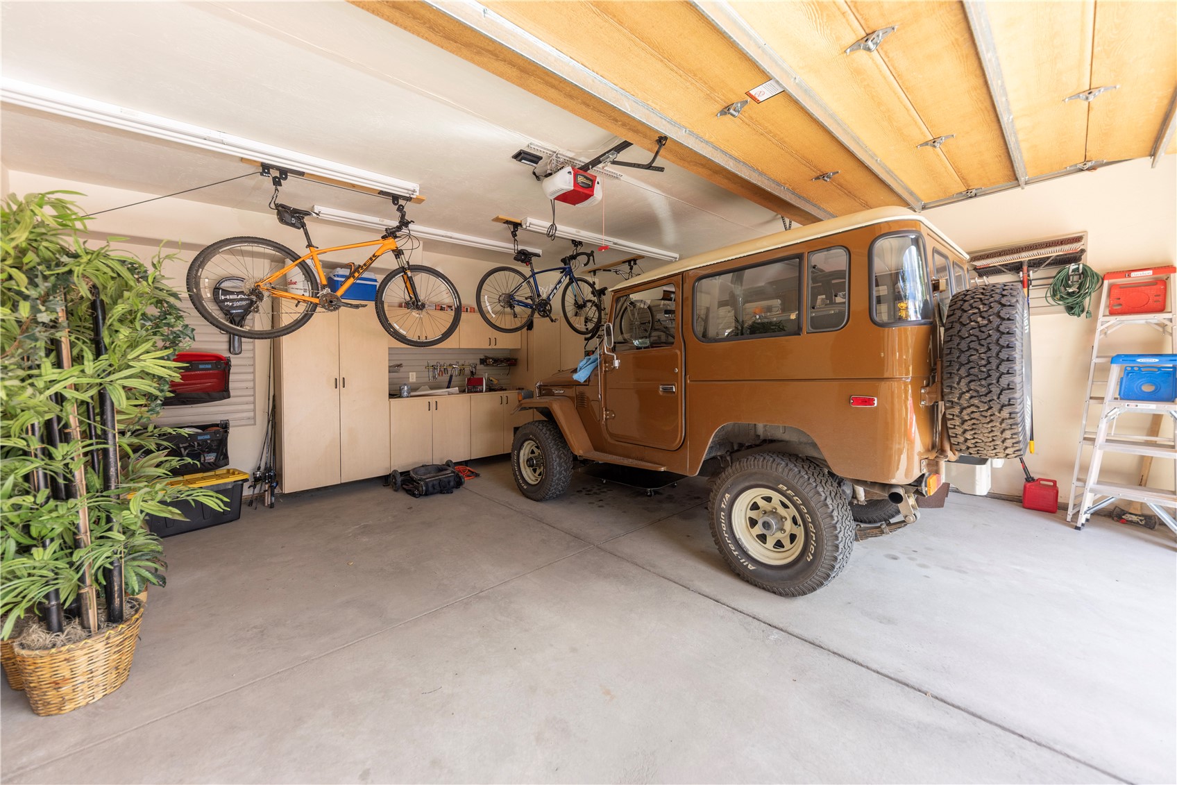 This spacious, well-organized garage features a classic brown vintage SUV parked prominently on the concrete floor. The space is equipped with light-toned cabinetry for storage, overhead bike racks holding two bicycles, and a tall step ladder tucked against the wall. A large potted plant adds a touch of greenery to the corner, creating a functional and tidy workshop environment.