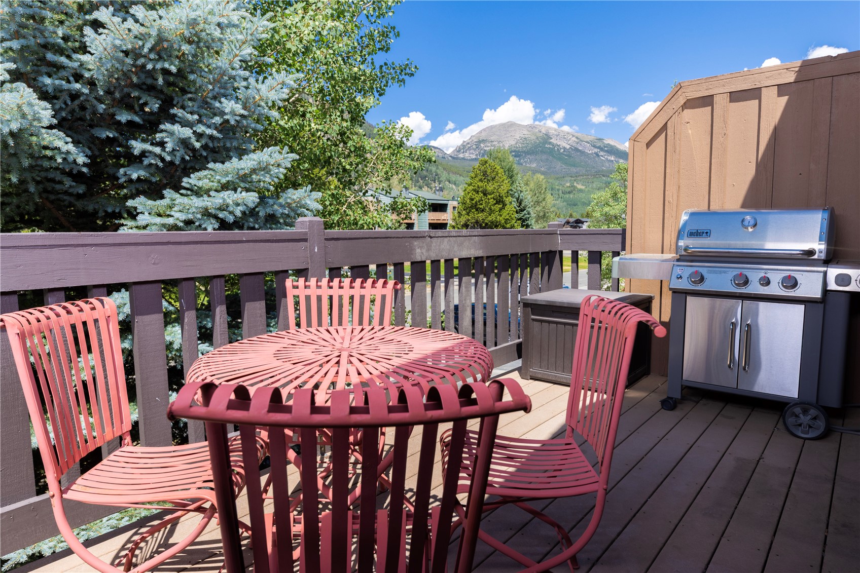 This elevated wooden deck offers a scenic outdoor living space complete with a metal bistro set and a stainless steel grill. The deck is enclosed by a simple wooden railing and provides a clear, picturesque view of the surrounding mountains and lush evergreen trees. The setting is bright and inviting, perfect for outdoor dining or relaxing while enjoying the natural landscape.