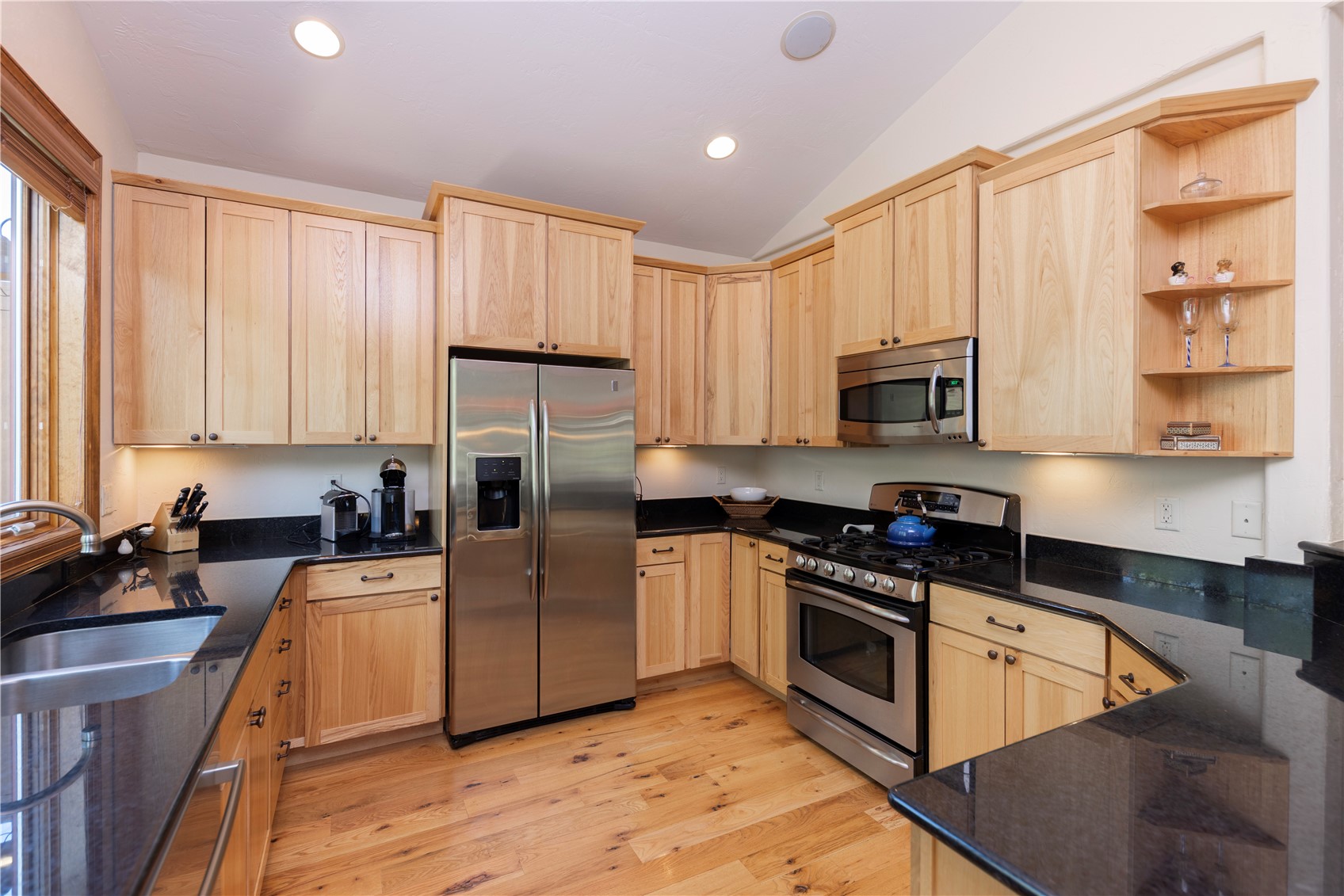 This kitchen features light wood cabinetry paired with dark stone countertops and stainless steel appliances, including a side-by-side refrigerator and a gas range. The space is illuminated by recessed lighting and under-cabinet lights, highlighting the warm tones of the hardwood flooring. An open shelving unit on the right adds a decorative touch, while the layout provides a functional workspace with a double-basin sink positioned under a window.