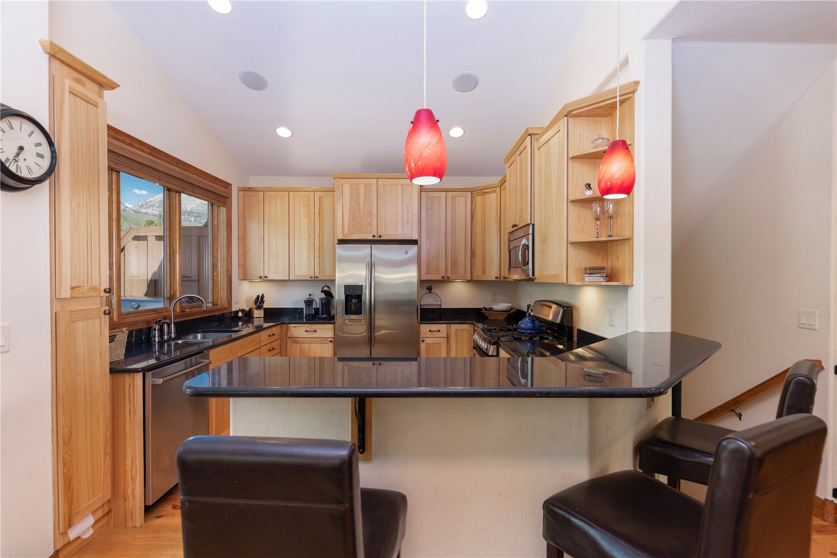 This kitchen features light wood cabinetry paired with dark stone countertops and stainless steel appliances. A breakfast bar with three dark leather stools provides casual seating, while two red pendant lights hang above the counter. The space is well-lit with recessed lighting and includes a window over the sink that offers a view of the outdoors.