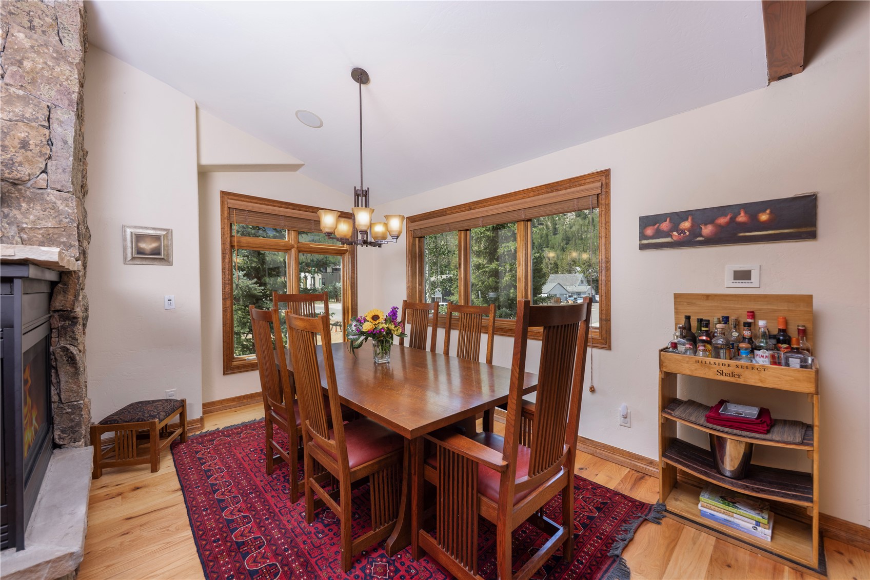 This dining room features a warm, traditional aesthetic with a large wooden table surrounded by six high-backed chairs. A vibrant, patterned area rug anchors the space, while a stone-clad chimney breast rises to the left, housing a fireplace. Large windows with wooden frames provide natural light and views of the outdoors, and a wooden bar cart stands to the right, completing the inviting, rustic atmosphere.