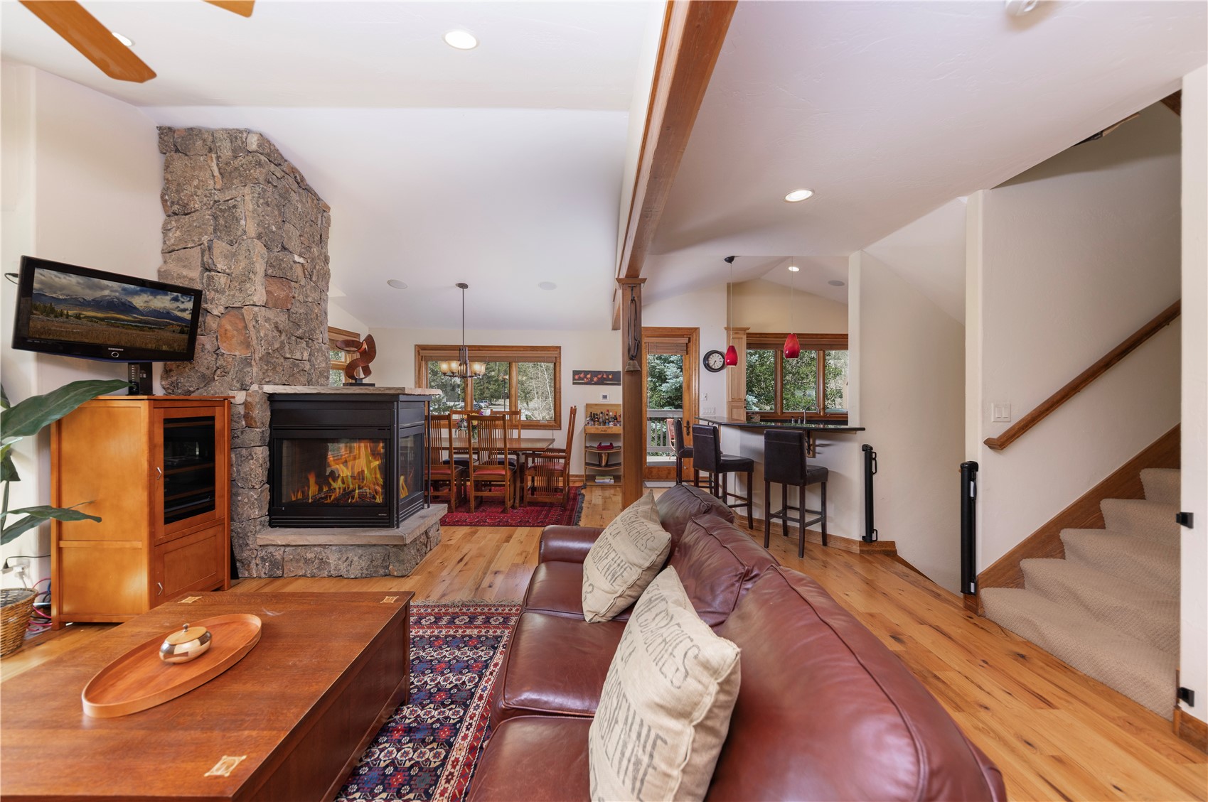 This open-concept living area features a prominent stone-clad chimney breast housing a fireplace, serving as the focal point of the room. A leather sofa sits on a patterned rug atop light wood flooring, while a dining area and a breakfast bar with pendant lighting are visible in the background. The space is connected to a staircase, creating a warm and inviting atmosphere with a rustic, mountain-lodge aesthetic.